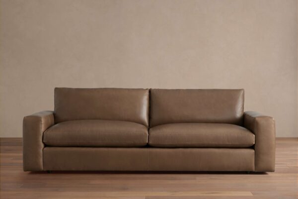Brown Kipton Wide Arm Leather Sofa with broad arms and two back cushions, placed on wood floor against a beige wall.