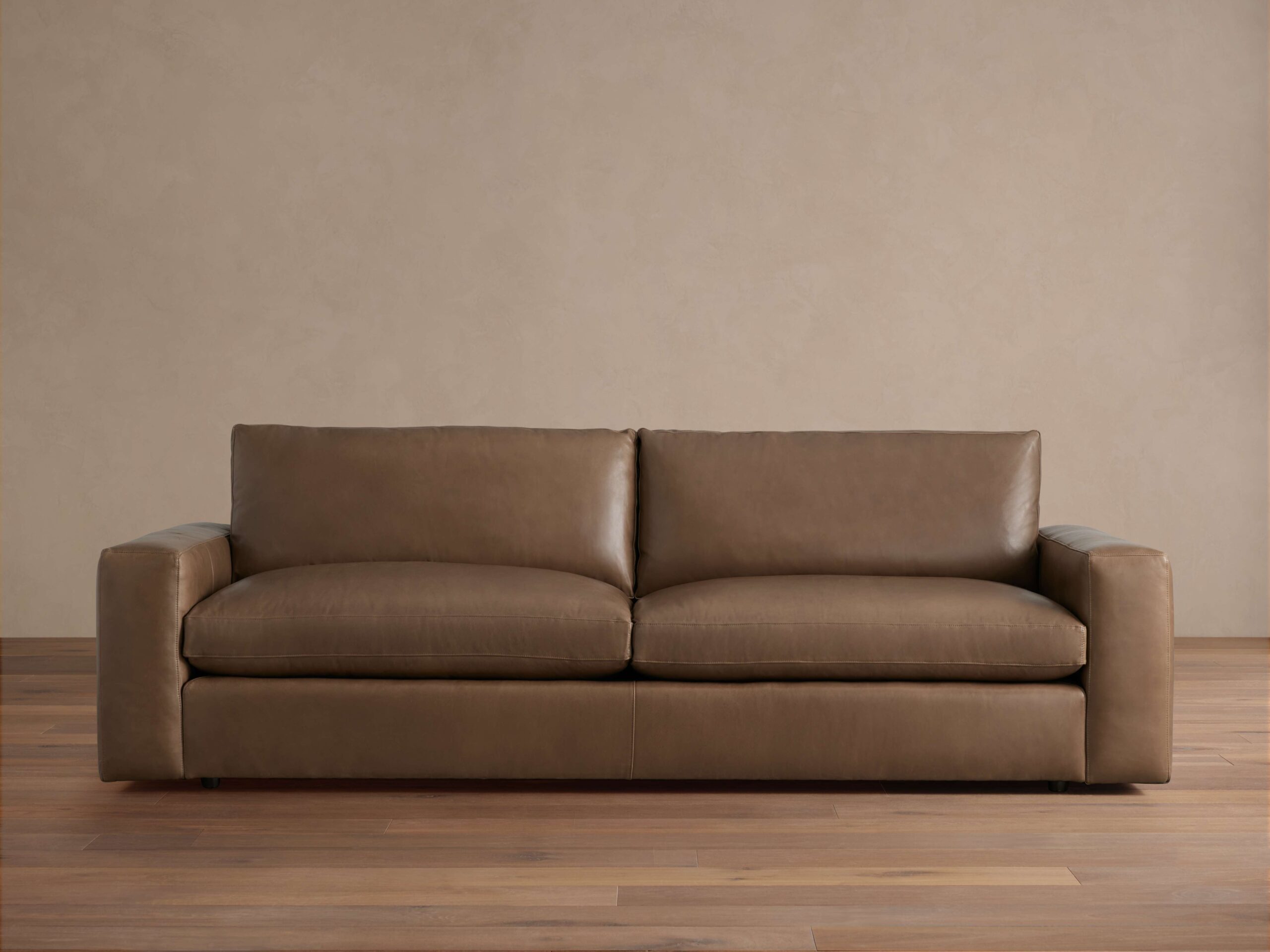 Brown Kipton Wide Arm Leather Sofa with broad arms and two back cushions, placed on wood floor against a beige wall.