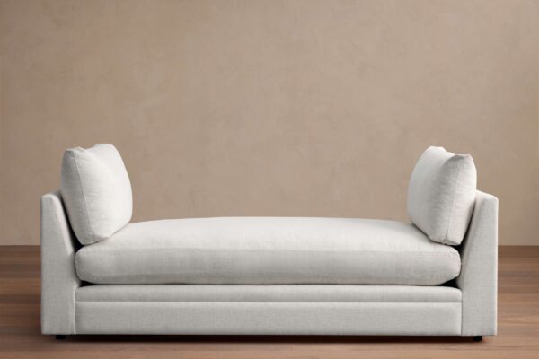 Cream-colored Pavo Daybed with plush cushions on either end, set against a neutral wall on a wooden floor.