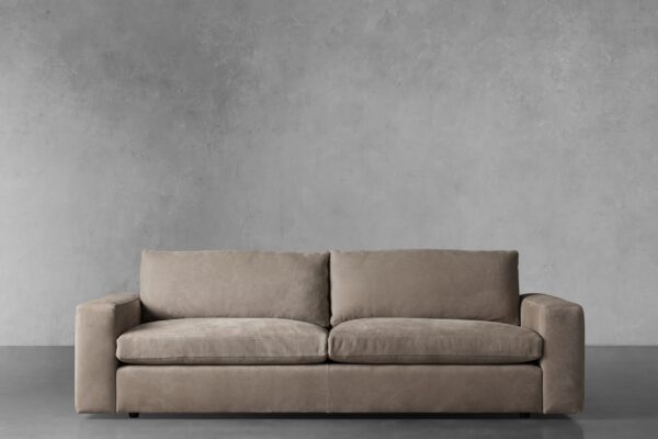 Kipton Wide Arm Nubuck Leather Sofa in beige, featuring wide arms and plush cushions, set against a gray concrete wall.